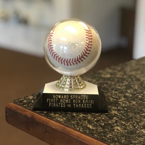 May include: A baseball trophy with a clear display case featuring a baseball signed "Rawlings". The base of the trophy is black with gold lettering that reads "Howard Sprague First Home Run 6/1/21 Pirates vs Yankees".