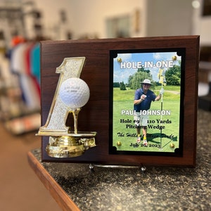 May include: A wooden plaque with a gold-colored number one and a golf ball on top. The plaque has a smaller plaque with a photo of a golfer and the text "HOLE-IN-ONE", "PAUL JOHNSON", "Hole #9 110 Yards", "Pitching Wedge", "The Hills at Mesa Verde", and "July 26, 2023".