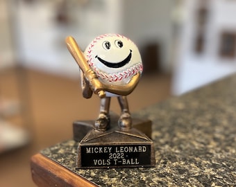 May include: A gold baseball trophy with a smiling baseball character holding a bat. The base of the trophy has a black plaque with the text "MICKEY LEONARD 2022 - VOLS T-BALL".