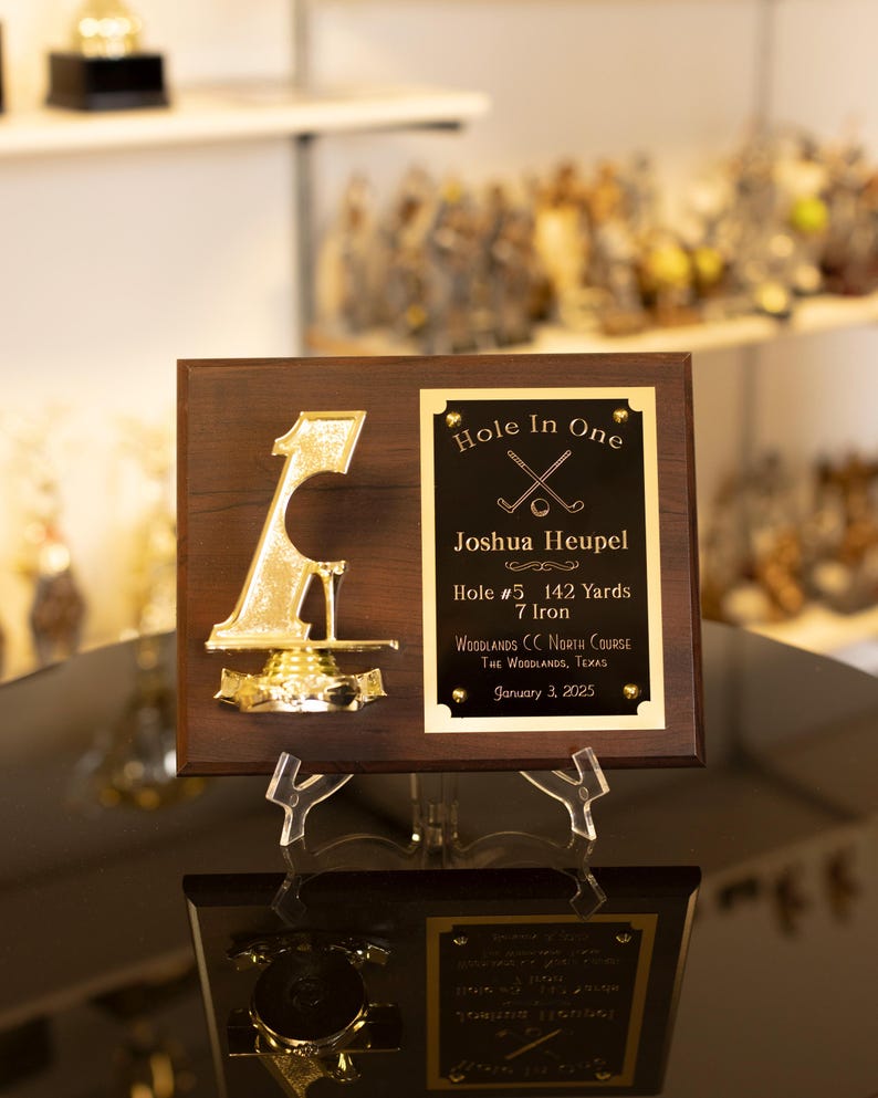 May include: A wooden plaque commemorating a golf "Hole in One." The plaque features a gold-colored "1" and a black plate with the text "Hole in One" and player details. The text includes "142 Yards" and the date "January 3, 2025."