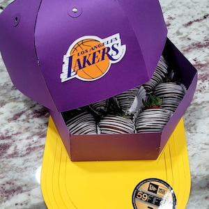 May include: A purple and yellow baseball cap shaped box filled with chocolate covered strawberries. The box has the Los Angeles Lakers logo on the front. The box is sitting on a white surface.