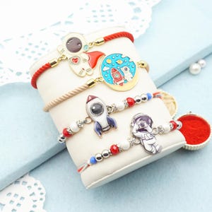 May include: Four bracelets with space-themed charms. One bracelet has a red cord with an astronaut charm. Another has a blue and red charm with a rocket and planet. The other two bracelets have white, red, and blue beads with rocket and astronaut charms.