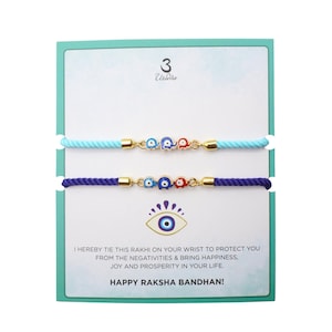 May include: Two Rakhi bracelets on a display card. The top bracelet has a light blue cord, the bottom a dark blue cord. Both feature gold-tone metal links with three small, colourful elephant charms. The card displays an eye icon and text.