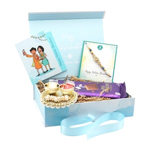 May include: A light blue gift box with a light blue ribbon, containing a chocolate bar, greeting cards, and a decorative tray. The cards feature illustrations and the text "Happy Raksha Bandhan".