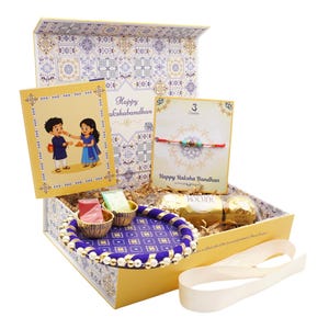 May include: A gift box set for Raksha Bandhan featuring a decorative purple and gold plate, a red thread bracelet on a card, a greeting card with an illustration of two children, two small containers of colored powder, and gold-wrapped chocolates.
