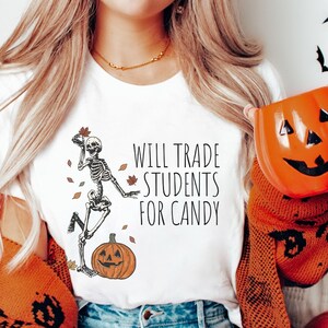 Pode incluir: Camiseta branca com um esqueleto segurando uma folha e uma ab&oacute;bora. O texto na camiseta diz "Will Trade Students For Candy".