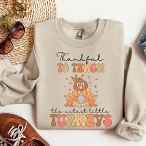 May include: Beige sweatshirt with a Thanksgiving-themed design. It features a cartoon turkey with the text "Thankful to Teach the cutest little TURKEYS".