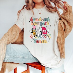 May include: A white t-shirt with the text "Student Support Team" in colorful retro letters. A cartoon pencil and book are holding hands and smiling.