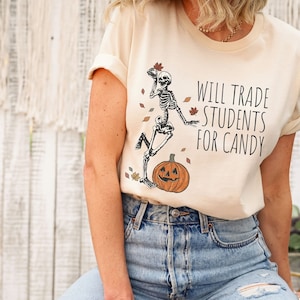 Pode incluir: Uma camiseta bege com um esqueleto segurando uma ab&oacute;bora e folhas. O texto na camiseta diz "Will Trade Students For Candy".