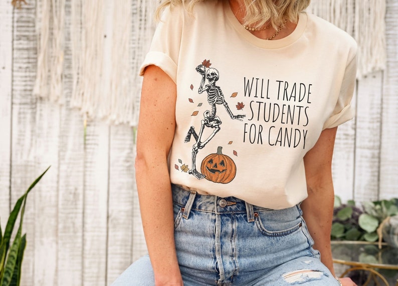 Pode incluir: Uma camiseta bege com um esqueleto segurando uma ab&oacute;bora e folhas. O texto na camiseta diz "Will Trade Students For Candy".
