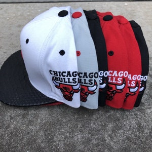 May include: A collection of Chicago Bulls baseball caps in various colors, including white, gray, black, and red. Each cap features the team's logo and the city name embroidered on the front. The caps have a black brim and a button on top.