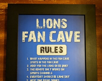 May include: A framed print with a blue background and white text that reads "Lions Fan Cave Rules". The rules are listed below: 1. What happens in the fan cave stays in the fan cave. 2. Root for the Lions or be quiet. 3. The remote only works on sports channels. 4. Everyday should be game day. 5. Next time bring drinks. 6. If you forget drinks bring food. 7. Always root against the Packers. My Lions. My Rules.