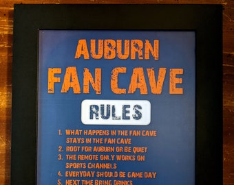 Può includere: Una stampa incorniciata con sfondo blu e testo arancione che recita "Auburn Fan Cave Rules". Le regole sono elencate di seguito: 1. Ciò che accade nella fan cave rimane nella fan cave. 2. Tifa per Auburn o stai zitto. 3. Il telecomando funziona solo sui canali sportivi. 4. Ogni giorno dovrebbe essere un giorno di partita. 5. La prossima volta porta le bevande. 6. Se dimentichi le bevande porta cibo. 7. Tifa sempre contro l'Alabama. La mia squadra. Le mie regole.