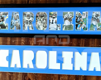 Könnte beinhalten: Zwei gerahmte Schilder mit dem Wort "CAROLINA" in weißen Buchstaben. Das obere Schild hat einen blauen Hintergrund und zeigt Fotos von Carolina Panthers Footballspielern in jedem Buchstaben. Das untere Schild hat einen blauen Hintergrund mit weißen Buchstaben.