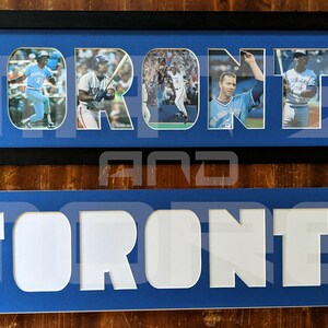 May include: A framed photo collage of Toronto Blue Jays baseball players. The collage is made up of individual photos of players in different poses, all set against a blue background. The word "TORONTO" is spelled out in white letters, with each letter containing a photo.