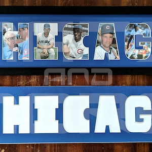 May include: A framed photo collage of Chicago Cubs baseball players. The word "CHICAGO" is spelled out with each letter containing a different player's photo. The frame is black with a blue mat. Below the frame is a blue sign with the word "CHICAGO" cut out in white.