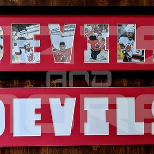May include: A framed black and white photo collage of hockey players with the word "DEVILS" spelled out in red with white letters. The photo collage is framed in black and is sitting on a wooden surface. Below the photo collage is a red sign with the word "DEVILS" spelled out in white letters.