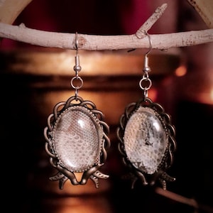 May include: A pair of antique-style earrings with oval-shaped, clear glass inserts showcasing a textured, off-white pattern. The earrings have a bronze-coloured, ornate frame and are suspended from silver-coloured hooks, displayed on a wooden branch.