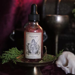 Puede incluir: Una botella de vidrio transparente de Aceite Ritual Perséfone con un cuentagotas negro. La etiqueta presenta una ilustración en blanco y negro de una mujer y las palabras "PERSEPHONE RITUAL OIL". La botella se encuentra sobre un pequeño pedestal dorado ornamentado.