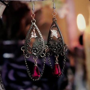May include: Ornate, antique-style earrings with a teardrop design. The earrings are bronze-colored with intricate filigree patterns, small white figures, and dangling red beads. They hang from a branch, with a blurred background.