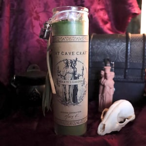 May include: A tall, green candle in a glass jar with a label that reads "Cat Cave Craft Hekate's Garden." The candle has a small, hanging vial and is decorated with dried leaves. Other items include a skull and a small statue.