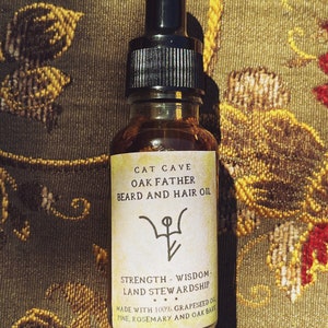 Op de afbeelding: Een klein glazen flesje met een druppelaar, gevuld met een lichtbruine vloeistof. Het etiket vermeldt "Cat Cave Oak Father Beard and Hair Oil" en bevat een symbool en de tekst "Strength - Wisdom - Land Stewardship. Made with 100% Grapeseed Oil, Pine, Rosemary and Oak Bark."