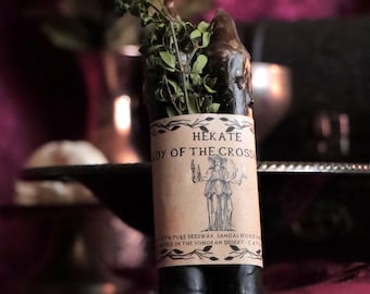 Puede incluir: Una vela oscura y cilíndrica con una etiqueta que dice "Hekate Lady of the Cross". La vela está adornada con vegetación seca y se encuentra sobre un fondo oscuro y borroso. La etiqueta presenta una ilustración de dos figuras.