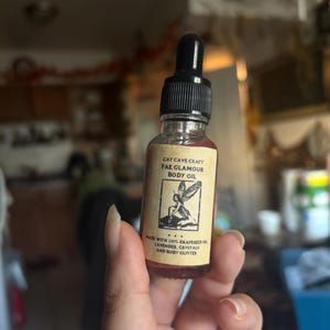 Może przedstawiać: Mała szklana butelka olejku do ciała Fae Glamour firmy Cat Cave Craft. Butelka ma czarną zakrętkę z kroplomierzem i etykietę z tekstem i ilustracją wróżki. Na etykiecie napisano, że olejek jest zrobiony z oleju z pestek winogron, lawendy, kryształów i brokatu do ciała.