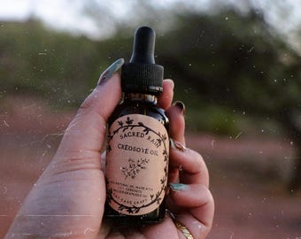 Può includere: Un flacone contagocce in vetro ambrato scuro con tappo nero e un'etichetta ovale color cuoio. L'etichetta presenta un bordo circolare a vite, una piccola illustrazione di una pianta e il testo Sacred Rain Creosote Oil, 100% natural oil, made with wild creosote, infused grapeseed oil, Cat Cave Craft.
