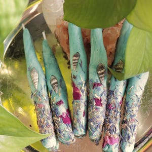 May include: A collection of teal-colored, cone-shaped candles decorated with dried flowers, seashells, and small decorative elements. The candles are arranged on a silver platter, with green leaves in the foreground.