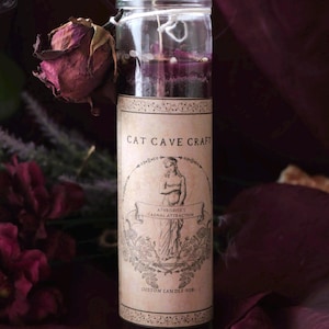 Könnte beinhalten: Eine Glas-Kerze mit lila Wachs und einem Etikett mit der Aufschrift "Cat Cave Craft". Das Etikett zeigt eine Illustration einer Frau und die Worte "Aphrodite's Carnal Attraction". Eine getrocknete Rose ist an der Kerze befestigt. Die Kerze ist in cm.