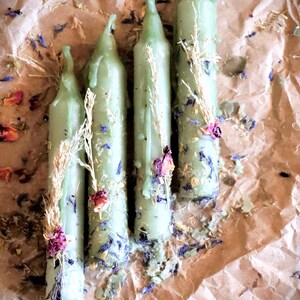 May include: Four light green candles of varying heights, adorned with dried flowers and herbs, rest on a brown paper surface. The candles have a dripped wax appearance and are decorated with small purple and blue floral accents.