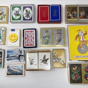 May include: Collection of vintage playing card decks, featuring various designs. Includes decks with floral, bird, and landscape illustrations, a smiley face, and a clown. Some decks are in boxes, others in clear plastic cases.