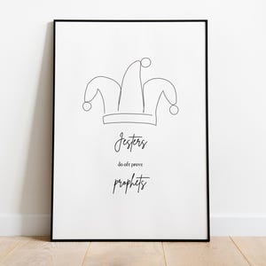 May include: A framed art print featuring a minimalist line drawing of a jester's hat. Below the hat, the words "Jesters do oft prove prophets" are written in a stylish script font. The artwork is set against a white background.