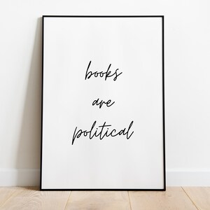 以下が含まれることがあります： 「books are political」という言葉が黒の優雅な筆記体で書かれた額入りのプリント。テキストは白い背景の中央に配置され、黒いフレームに収められています。プリントは、木製の床に白い壁に立てかけられています。