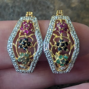 May include: A pair of gold-toned earrings with a floral design. Each earring features a central floral arrangement with red, black, and green gemstones, surrounded by a border of small, clear stones. The earrings have a rectangular shape with a decorative top.