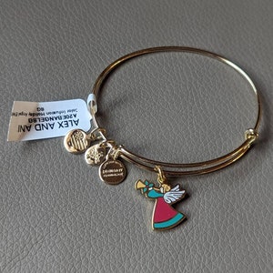 Op de afbeelding: Een gouden armband met een bedel van een engel die trompet speelt. De bedel is rood, blauw en goud. De armband ligt op een grijs oppervlak. De tekst op het etiket luidt "ALEX AND ANI A20EBANGLE  Julee  Illumination Holiday Gift  SG"