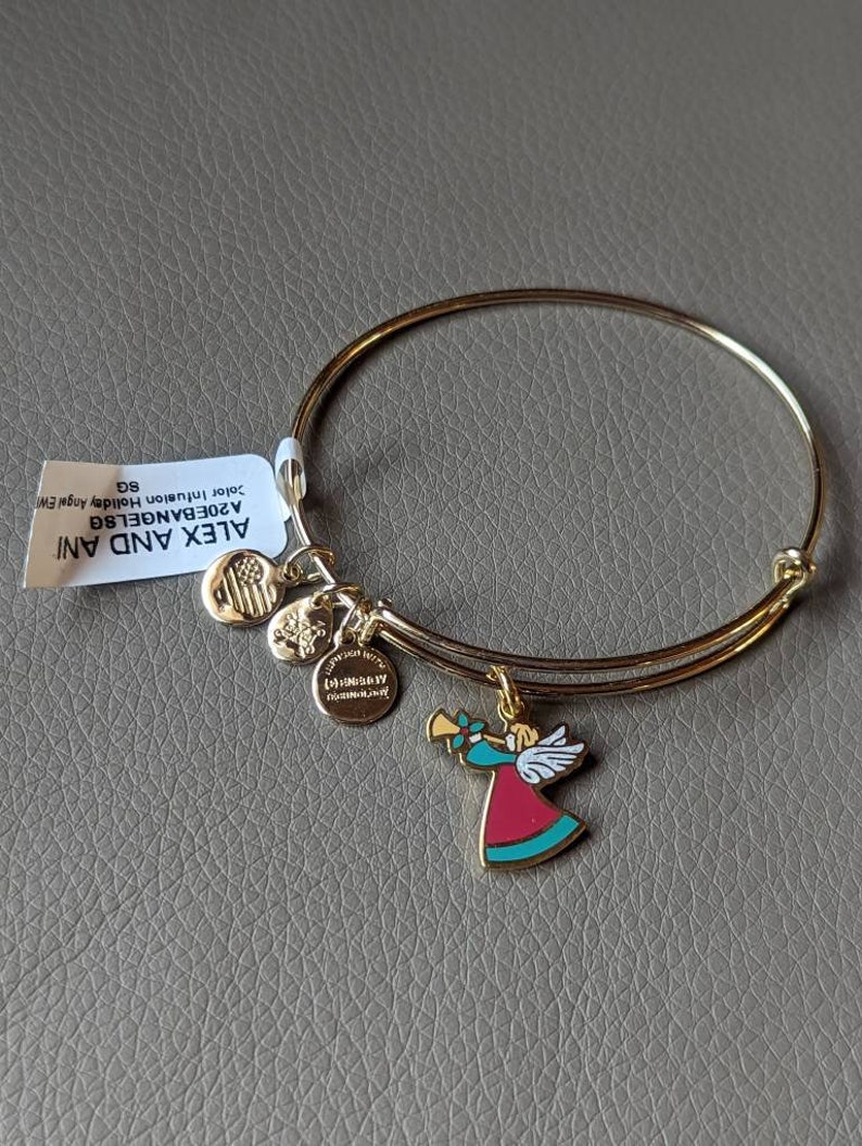 Op de afbeelding: Een gouden armband met een bedel van een engel die trompet speelt. De bedel is rood, blauw en goud. De armband ligt op een grijs oppervlak. De tekst op het etiket luidt "ALEX AND ANI A20EBANGLE  Julee  Illumination Holiday Gift  SG"