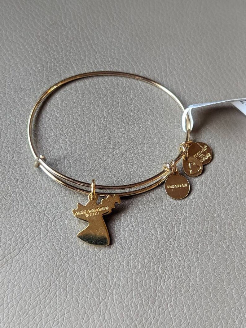 Op de afbeelding: Een gouden armband met een engelbehang. De hanger heeft de woorden "Angel Adornments" en "97101" erop gegraveerd. De armband heeft twee kleine hangers met de woorden "Alex and Ani" en "Find Your Wings" erop gegraveerd.