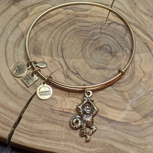 May include: A gold bangle bracelet with a monkey charm hanging from it. The bracelet has three charms attached to it, one with the words "Made with Energy Technology", one with a heart and an American flag, and one with the word "Alex and Ani".