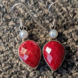 May include: A pair of silver earrings featuring teardrop-shaped, faceted red gemstones. Each earring has a small, round, white pearl above the red stone and a silver hook.