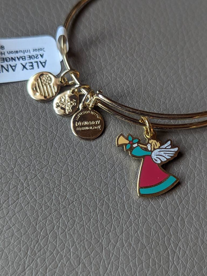 Op de afbeelding: Gouden armband met drie bedels. De bedels zijn een kleine Amerikaanse vlag, een kleine bedel met een hart en de woorden "Ik hou van je" en een grotere bedel van een engel die trompet speelt. De engel draagt een rode en turquoise jurk met witte vleugels.