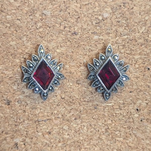 May include: A pair of silver-toned stud earrings. Each earring features a central, deep red, diamond-shaped stone, surrounded by a decorative, silver-toned frame with small, round accents. The earrings are set against a textured, brown background.