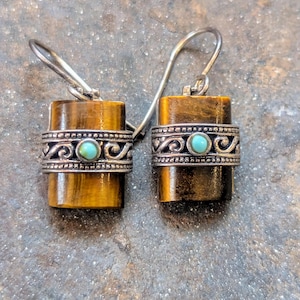 May include: A pair of handcrafted earrings featuring rectangular tiger's eye stones. Each earring has a decorative silver band with an ornate design and a small turquoise stone. The earrings have silver ear wires.