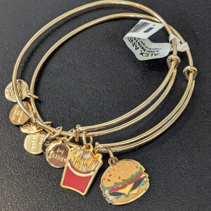 May include: Gold-tone bangle bracelets with food-themed charms. Charms include a hamburger, french fries, and round charms with text. The bracelets are on a dark surface.