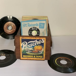 Pode incluir: Uma caixa de madeira com um design de pêssego e o texto "Peaches Records & Tapes" está cheia de discos de vinil antigos. Os discos são etiquetados com os nomes "RCA Victor" e "Victor".