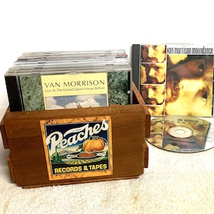 Może przedstawiać: Drewniana skrzynka z etykietą "Peaches RECORDS & TAPES" zawiera kilka płyt CD, w tym "Van Morrison Live At The Grand Opera House Belfast". Widoczna jest również płyta CD "Van Morrison Moondance". Skrzynka jest brązowa, a płyty CD są w przezroczystych etui.