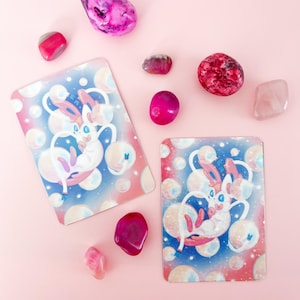 May include: Two rectangular cards with a cartoon character illustration, surrounded by pink, purple, and red gemstones. The cards feature a white and pink creature with blue accents, set against a blue and pink bubble background. The gemstones vary in size and shape.