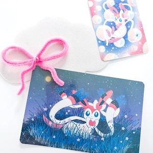 May include: Two illustrated cards featuring a Sylveon character, one with a starry night background and the other with bubbles. A pink pipe cleaner bow and a white cloud-shaped cutout are also present.