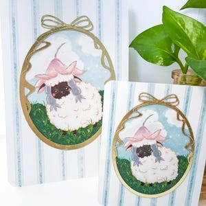 May include: Two decorative cards with a cartoon sheep wearing a pink flower hat. The sheep is inside a gold oval frame on a striped background. The cards are different sizes.